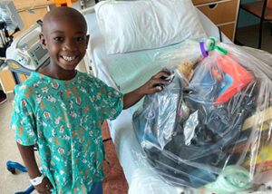 Kevin, a black boy, in his hospital gown standing next to a hospital bed with his wrapped bag of fun on the bed.