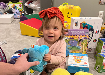 Ruth, a little white girl, sitting on the floor with toys all around her. She is holding a blue stuffed animal and another hand is holding the other end of the stuffed animal. We can't see the person. We only see the hand. Ruth is looking at the person behind the camera.
