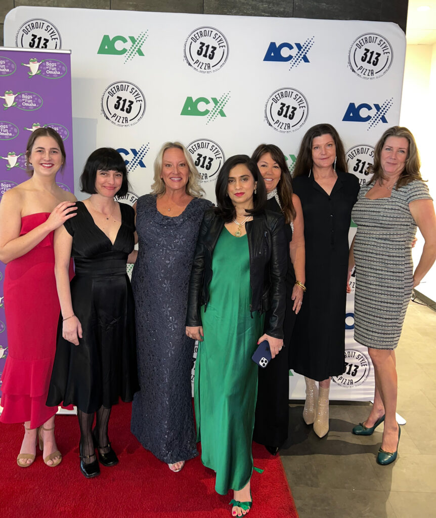 7 women dressed in evening gowns standing on a red carpet with two logo backdrops behind them. One is purple and has the Bags of Fun Omaha logo and the other larger one has the Detroit Style 313 Pizza and ACX logos. These logos are repeated in a pattern and the backdrop is white.