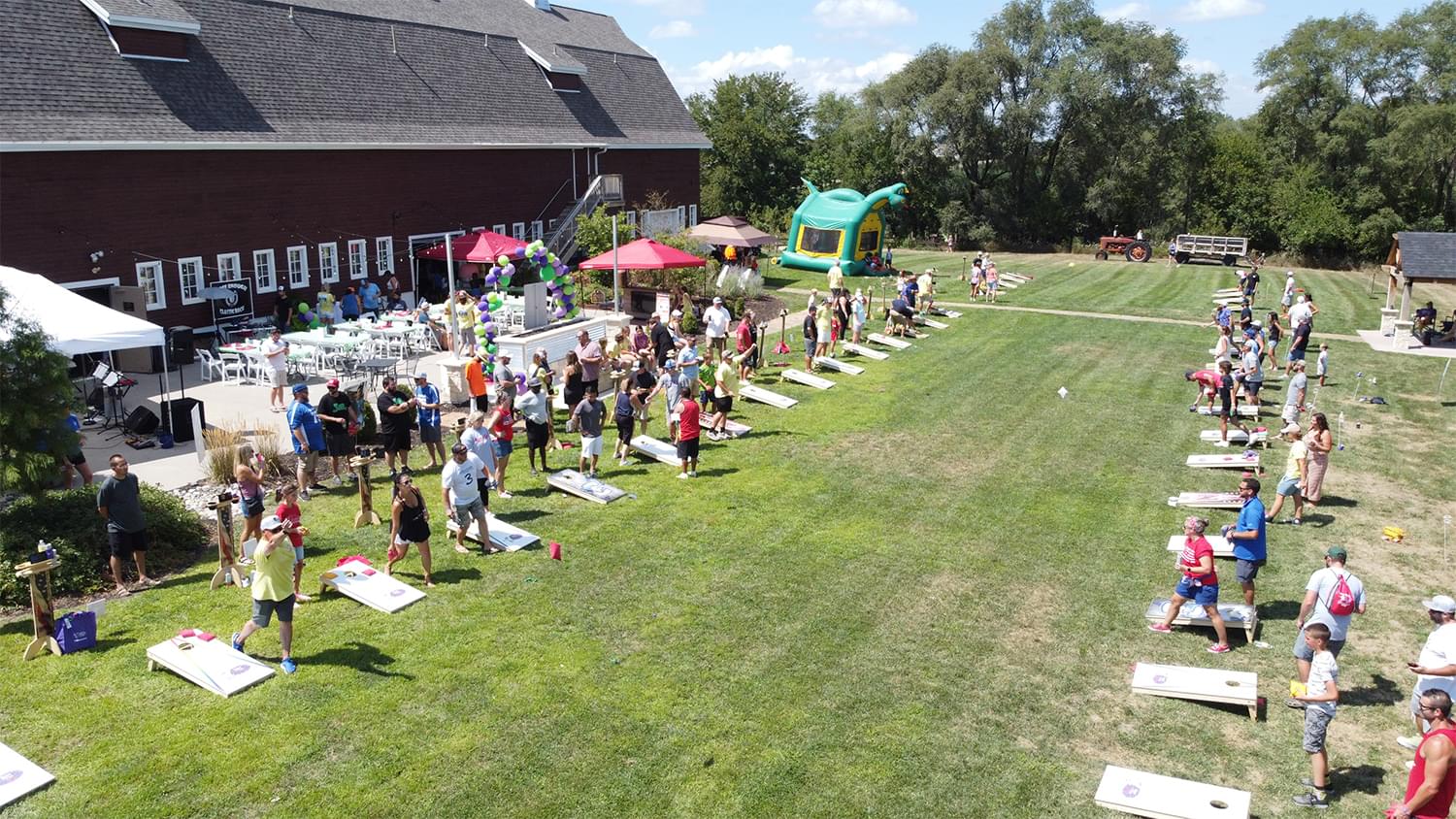 Drone view o the cornhole tournament. We see a large red barn in the background. In front of the bar there is a patio with chairs, a stage, and balloons. In front of that is a large grass area where there are rows of cornhole games lined up and people on either side playing cornhole. 