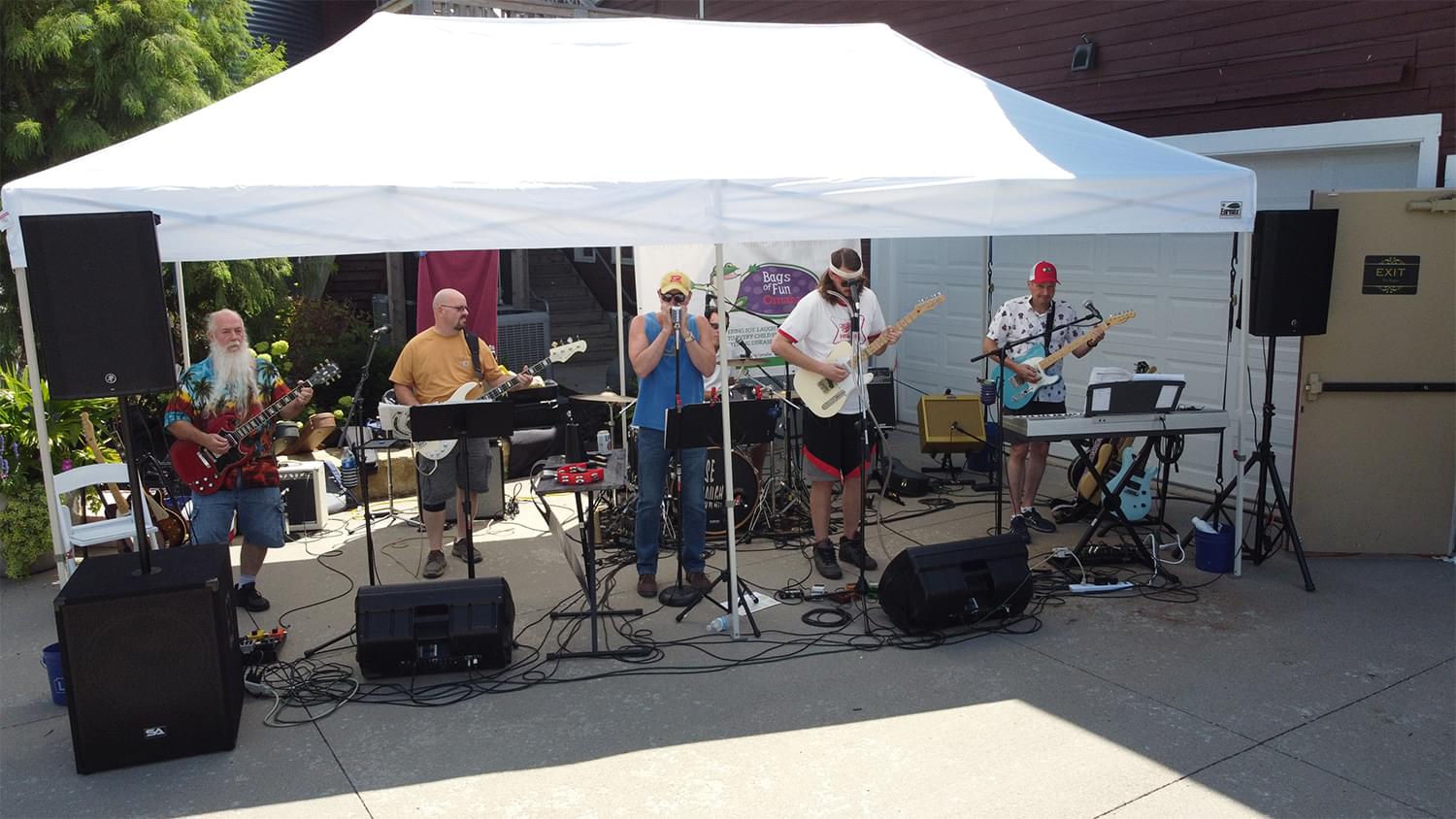 A 5 person band, 4 people playing guitars and one person playing the harmonica. They are under a large white tent and you see all the band equipment around them, including speakers, a keyboard, and drums.