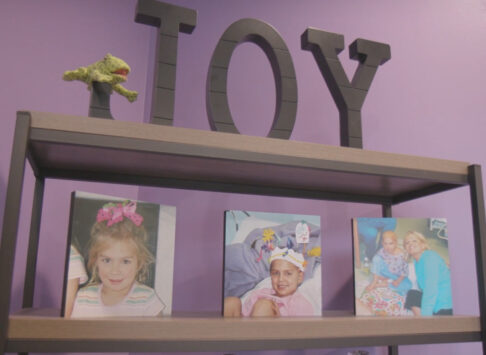 A close up of a book shelf with a purple background. At the top of the book shelf are the words, Joy, in metal. There is a toy frog resting on the loop of the J. On the second shelf are 3 pictures of Gaby.