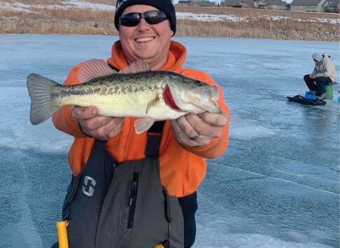 A man in an orange jacket and gray fishing overalls is holding up the fish he caught. He is standing on the ice.