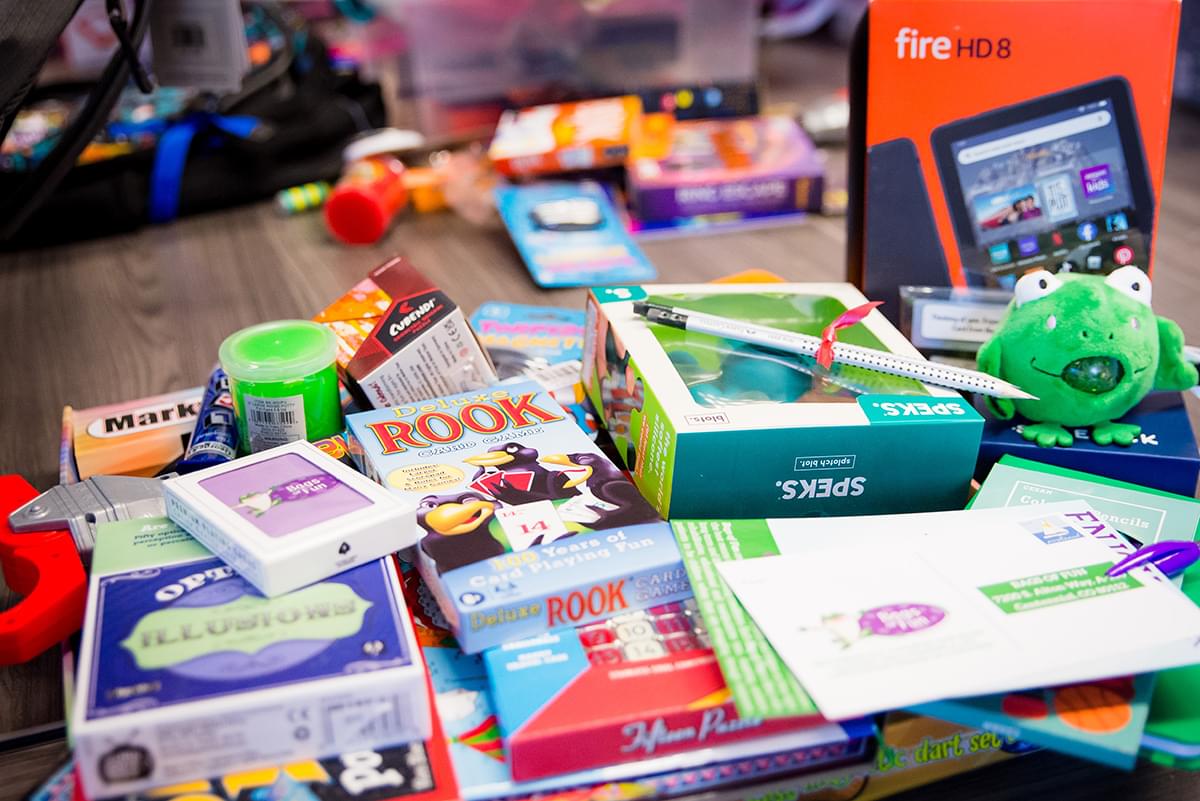 There is a large stack of toys sitting on a big wooden table. Some of the toys are blurred out but we can see a deck of Bags of Fun playing cards, the Fire HD 8 orange box, a stuffed green frog with beads that come out of its mouth, a Speks box, two pencils tied together with a red ribbon, a Rook, and other toys and games.
