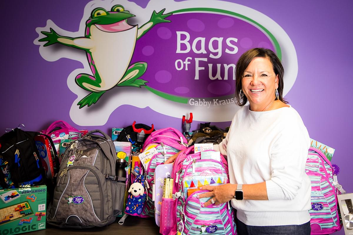 Tammy Krause is wearing a white sweater and jeans and is standing in front of a table full of bags. She is holding the pink, blue, and gray unevenly striped backpack and is smiling at us. There are lots of other bags on the table as well with toys next to or in them. The large Bags of Fun logo on a purple wall is behind them.