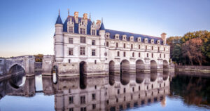 Loire Valley Chateau castle. The base of the castle is in the water. It looks like a bridge with archways forming the base of the castle. There is a beautiful reflection of the castle in the still water. The castle is white and gray with a blue roof and there are trees behind the castle with green and orangy read leaves, suggesting it is fall time.