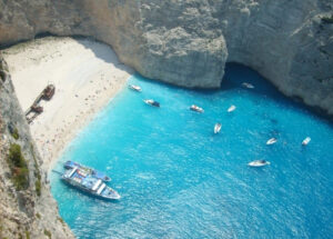 Overhead view of a gorgeous beach and pristine blue water and boats in Greece. We see large rock walls surrounding the beach and water, suggesting this is an inlet within a cliff or mountain formation.