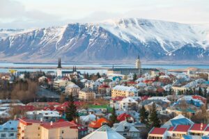 Iceland town with beautiful snow peeked mountains in the background and a body of water that is running between the mountains and the town. The town has lot of buildings, homes, and churches. We see lots of red, blue, yellow, and white, roofs and buildings.