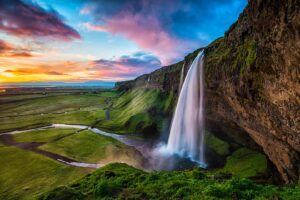 Iceland waterfall with a sunset in the distance. There are breathtaking blues, purples, and oranges in the sky and reflecting in the water. There is also greenery at the base and all the way up to the waterfall.