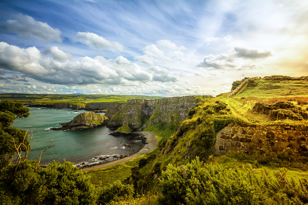 A beautiful Irish coastline with grassy green multilayered cliffs on the right and at the base towards the left there is water that meets the shore. The green cliff plains stretch far and wide in the background and there is a blue sky with lots of fluffy white clouds.
