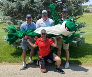 4 men and, Chuckles, the Bags of Fun mascot, posing for a picture on the golf course path with a tree behind them. One person in a red shirt is kneeling on one knee in the middle and has his hands up at his shoulders because he is helping to hold up chuckles, who is lying horizontally across the kneeling mans hands. There are 3 men behind Chuckles and they are all holding him up as well.