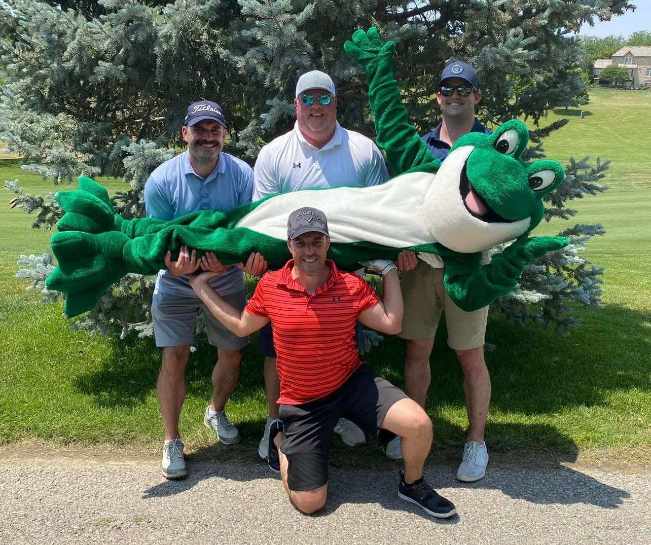 4 men and, Chuckles, the Bags of Fun mascot, posing for a picture on the golf course path with a tree behind them. One person in a red shirt is kneeling on one knee in the middle and has his hands up at his shoulders because he is helping to hold up chuckles, who is lying horizontally across the kneeling mans hands. There are 3 men behind Chuckles and they are all holding him up as well.