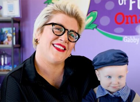 A mom with black glasses and a trendy short blond haircut that sticks up a bit. She has black thick rimmed glasses on, is smiling with bright red lipstick, and has her head tilted to one side a little. She is holding a 2 or 3 year old in her arms. He has blue eyes, is smiling at the camera, and looks very sophisticated with a blue beret hat and dark blue sweater with a blue and white checker shirt collar sticking out the top of the sweater. He has a little smile on his face as well. We can see the purple backgorund behind them with part of the Bags of Fun Omaha logo on the wall.