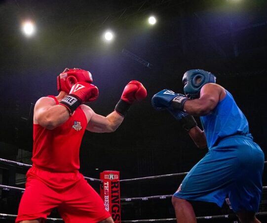 two men in a boxing ring with the bright lights above them. One man is wearing a red uniform and the other is wearing blue. They both have their fists up and appear to be in the middle of a boxing match.