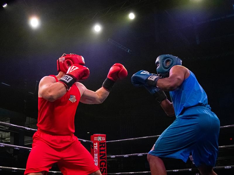 two men in a boxing ring with the bright lights above them. One man is wearing a red uniform and the other is wearing blue. They both have their fists up and appear to be in the middle of a boxing match.