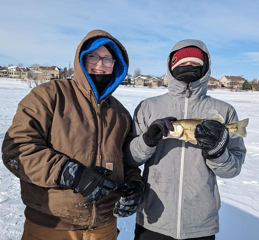 Two young adults all bundled up with jackets, winter hats, gloves, and masks outside. One is holding a small fish.