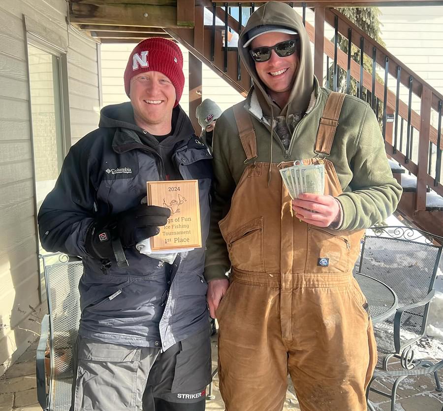 Two adults with snow gear on are standing together. One is holding their aware money and the other is holding their aware plaque