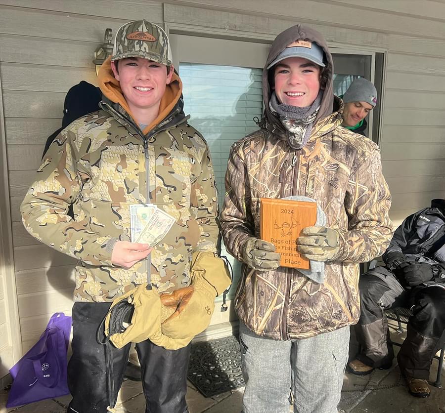 Two teenagers with camo jackets on are standing together. One is holding their award plaque and the other is holding their award money.