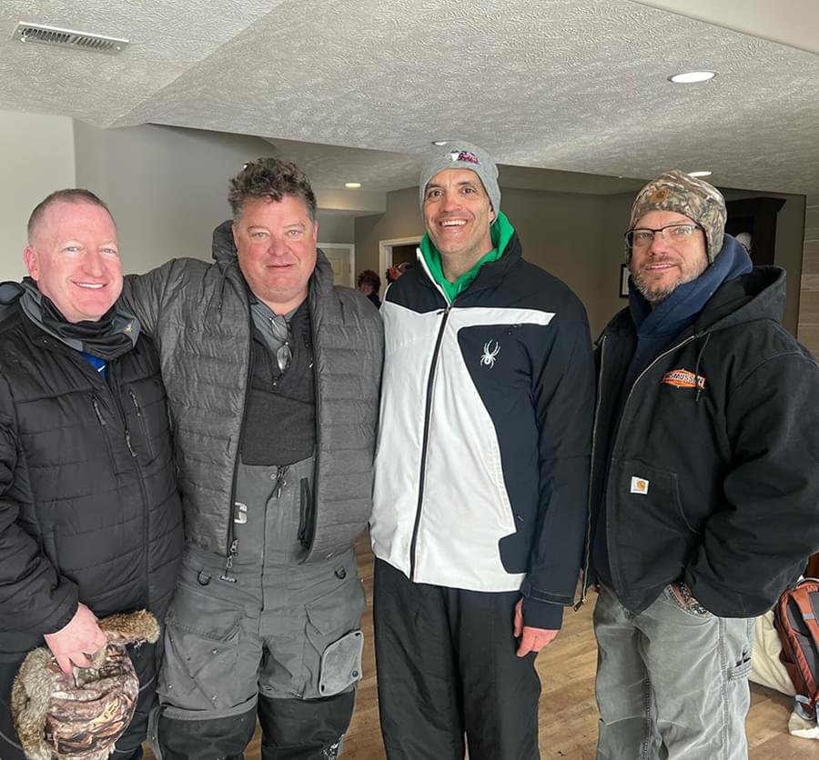 four men wearing winter clothes and standing inside. They are gathered together and smiling for the photo.