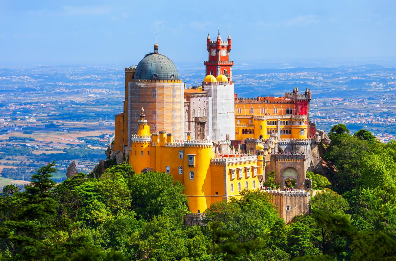 A huge orange, yellow, and red castle on top of a lush green hill with vast land behind it in the background.