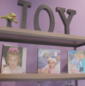 A close up of a book shelf with a purple background. At the top of the book shelf are the words, Joy, in metal. There is a toy frog resting on the loop of the J. On the second shelf are 3 pictures of Gaby.