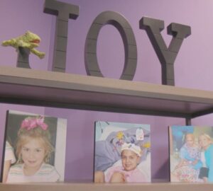 A close up of a book shelf with a purple background. At the top of the book shelf are the words, Joy, in metal. There is a toy frog resting on the loop of the J. On the second shelf are 3 pictures of Gaby.