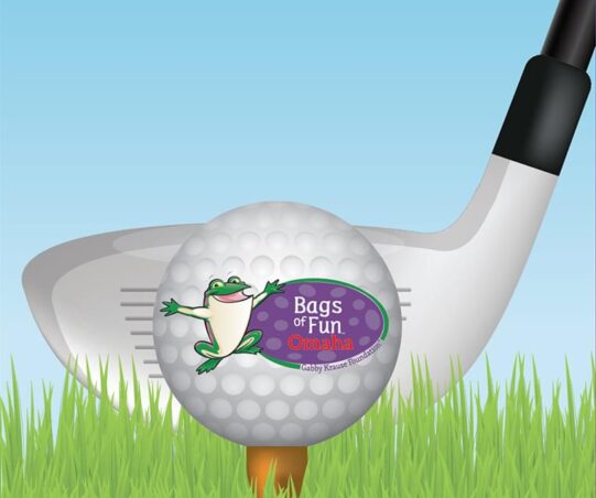 Illustration of a golf club with a golf ball in front in the grass on a tee. The ball has the Bags of Fun Omaha logo on it.