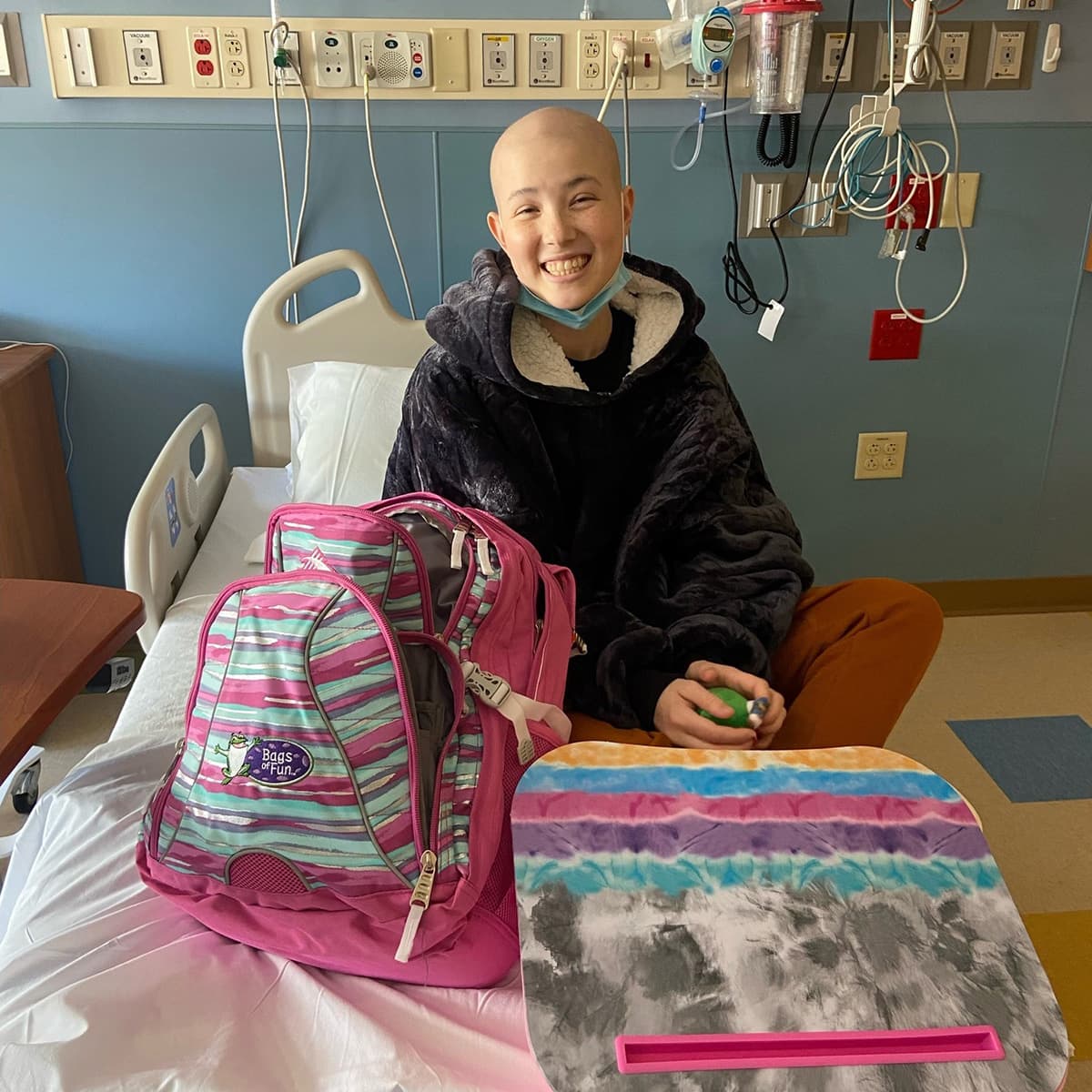 Teenager sitting in a hospital bed smiling with a fuzzy sweater and her bag of fun.