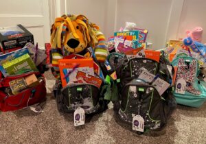 A group of backpacks stuffed with games, toys, and stuffed animals set down on the carpet.