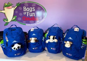 Four blue backpacks with a stuffed animal coming out of each front pocket and green water bottles in the side pockets. In front of a purple wall with the Bags of Fun Omaha logo.