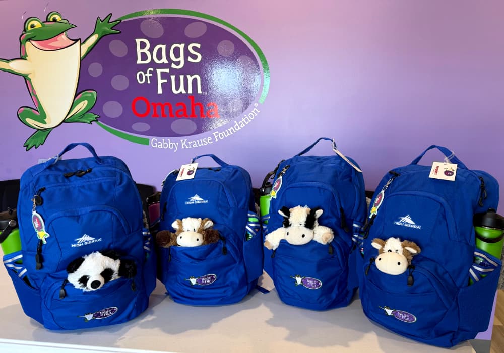 Four blue backpacks with a stuffed animal coming out of each front pocket and green water bottles in the side pockets. In front of a purple wall with the Bags of Fun Omaha logo.